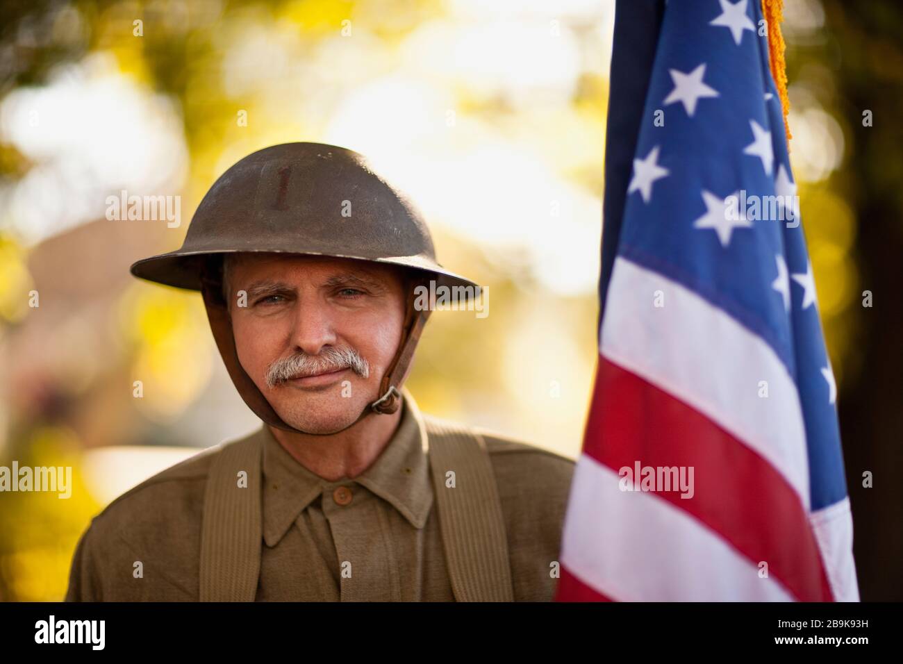 Reenactor maschile maturo che indossa un casco da combattimento in acciaio della Guerra Mondiale americana e uniforme e che tiene una bandiera americana pone per un ritratto. Foto Stock