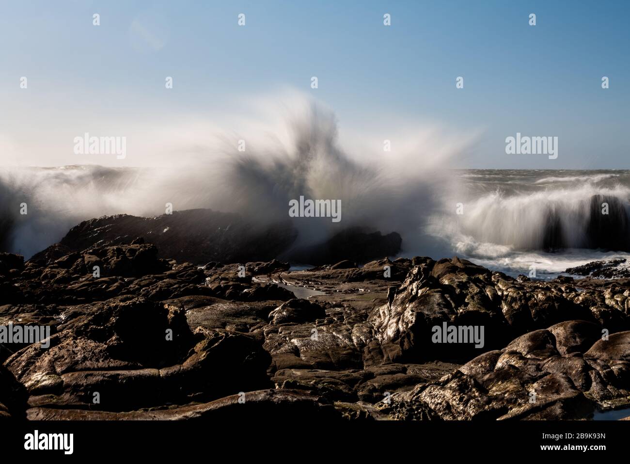 Lunga esposizione di onde che si infrangono sulle rocce costiere sotto il cielo blu Foto Stock