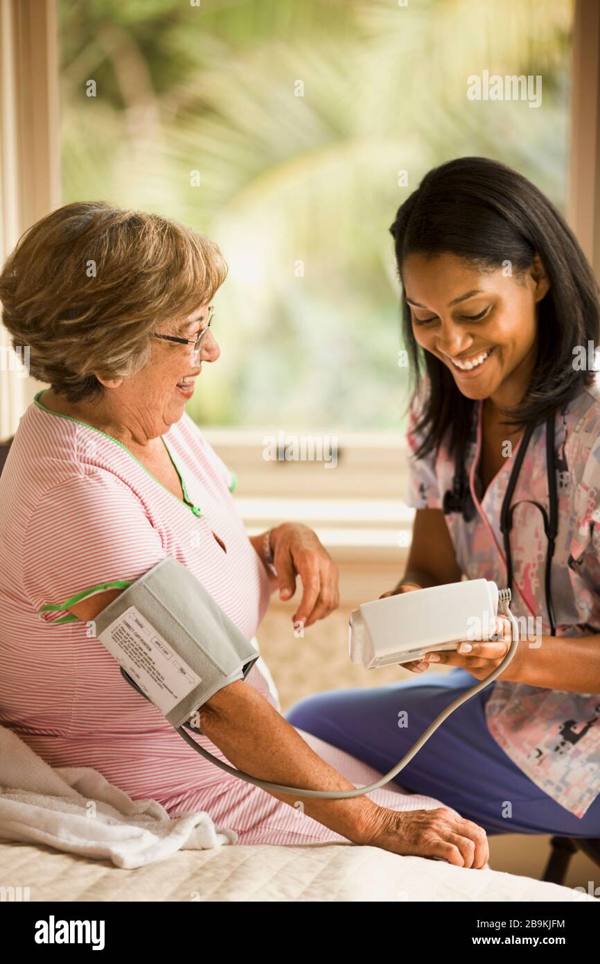 L'infermiera femminile utilizza un bracciale per la misurazione della pressione sanguigna e un monitor per la misurazione della pressione su una donna anziana che siede sul letto. Foto Stock
