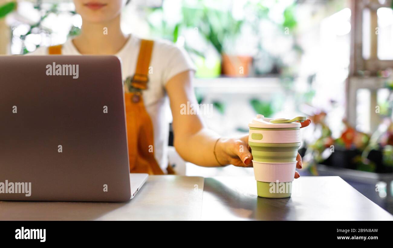Freelancer femminile in possesso di tazza di caffè/tè riutilizzabile, utilizzando il computer portatile, comunica su Internet in casa giardino / serra. Messa a fuoco selettiva. Lavoro da ufficio confortevole Foto Stock