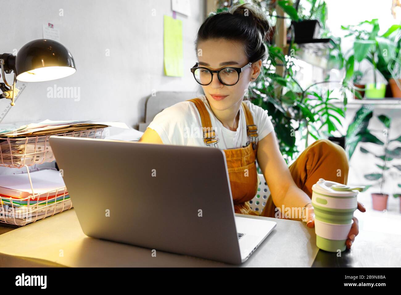Giovane donna giardiniere in bicchieri utilizzando il computer portatile, comunica su internet con il cliente in casa giardino / serra, tenere riutilizzabile caffè / tè tazza.Cozy off Foto Stock