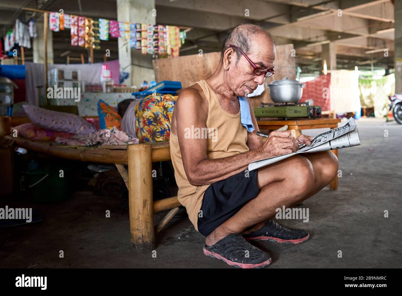 Uomo che fa i crosswords in un garage del mercato pubblico del carbonio in cui la gente vive dopo che hanno perso le loro case in un fuoco. Foto Stock