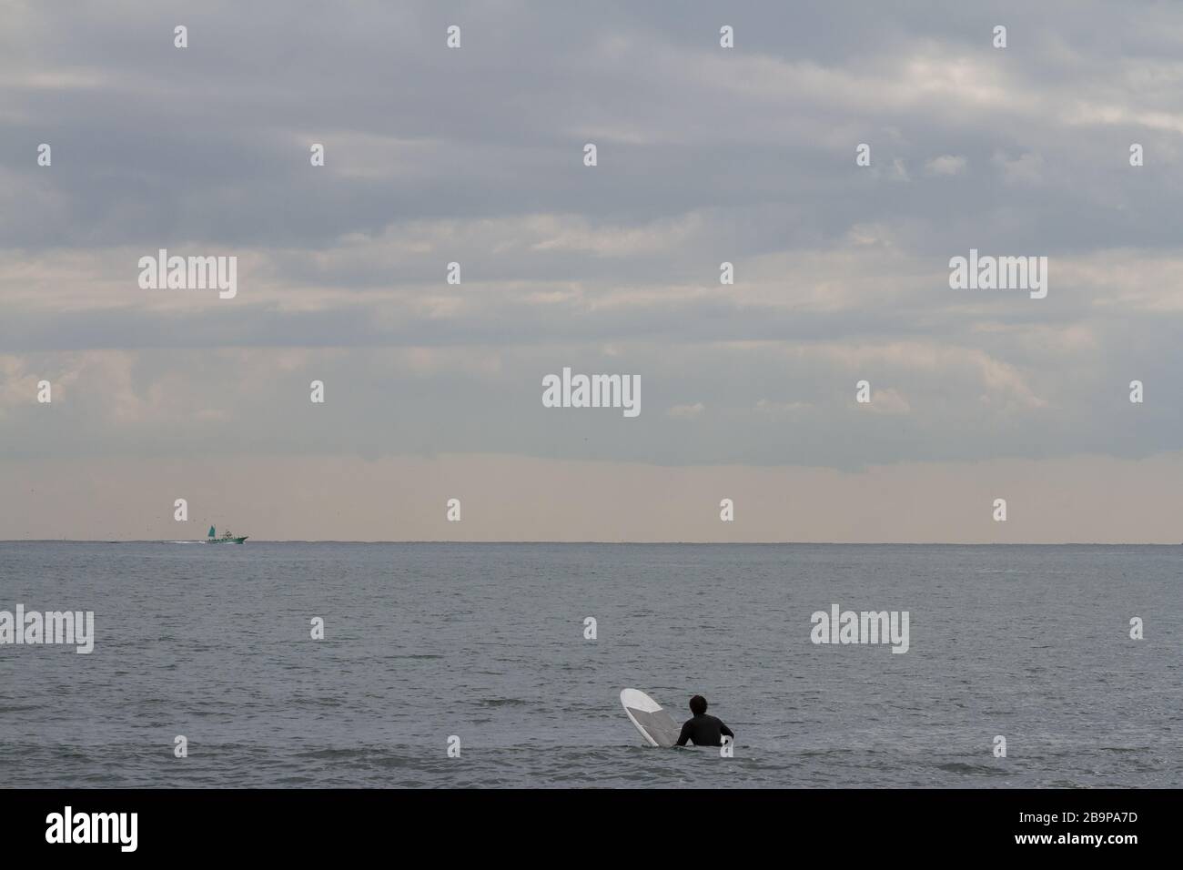 Un surfista si siede sulla sua tavola da surf in attesa di onde a Enoshima, mentre una barca da pesca passa all'orizzonte, Kanagawa, Giappone. Foto Stock