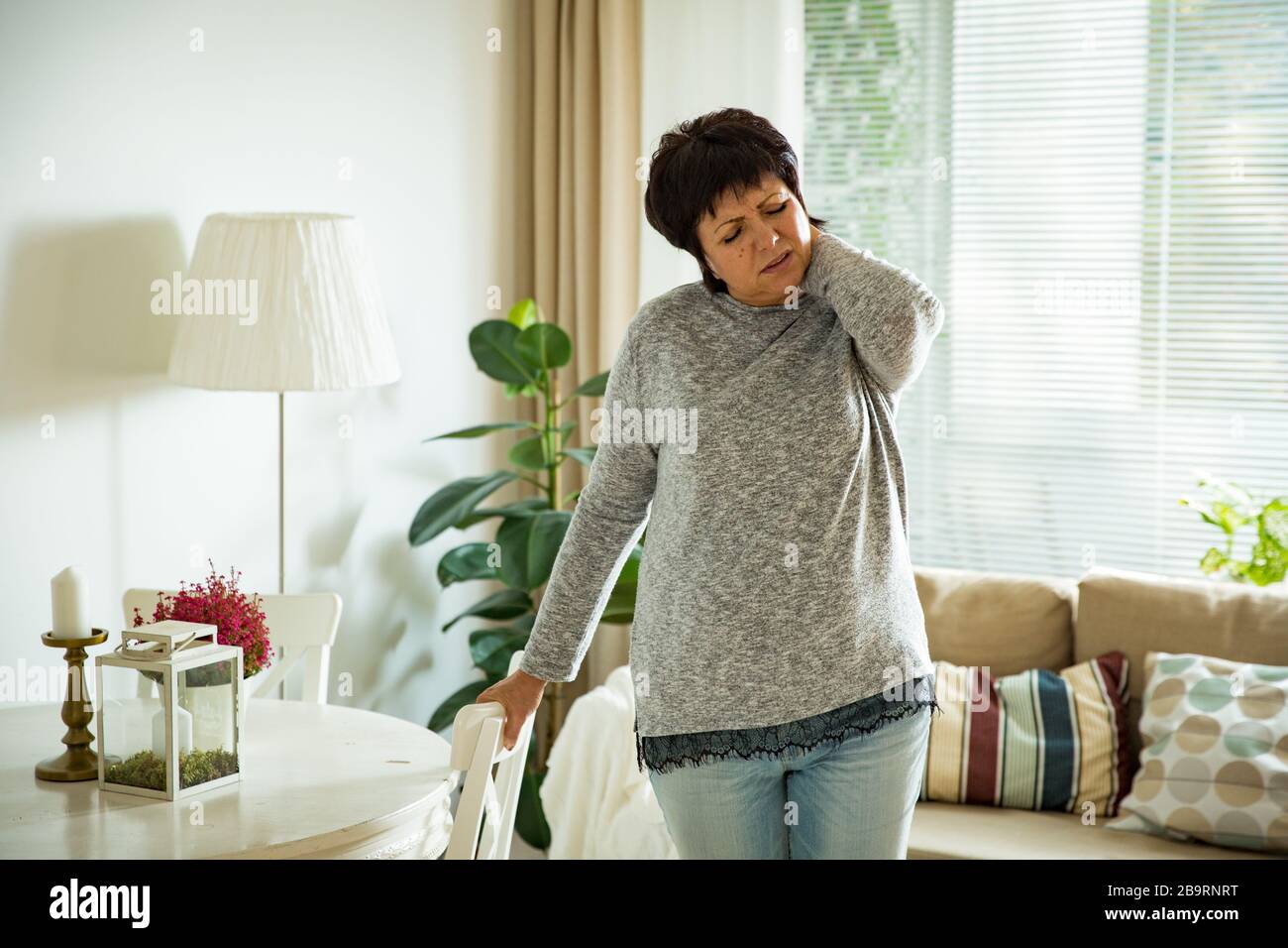 Donna matura che soffre di mal di schiena a casa. Massaggiando il collo con la mano, sentendosi esausto, stando in piedi nel soggiorno. Foto Stock