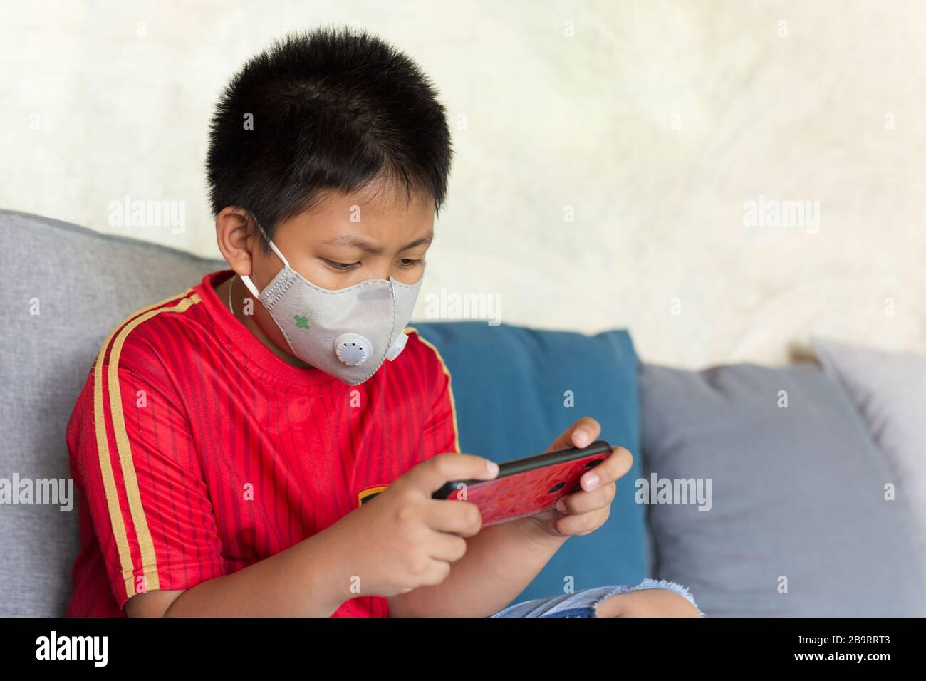 Ragazzo asiatico con maschera protettiva gioco sul telefono cellulare a casa. Foto Stock