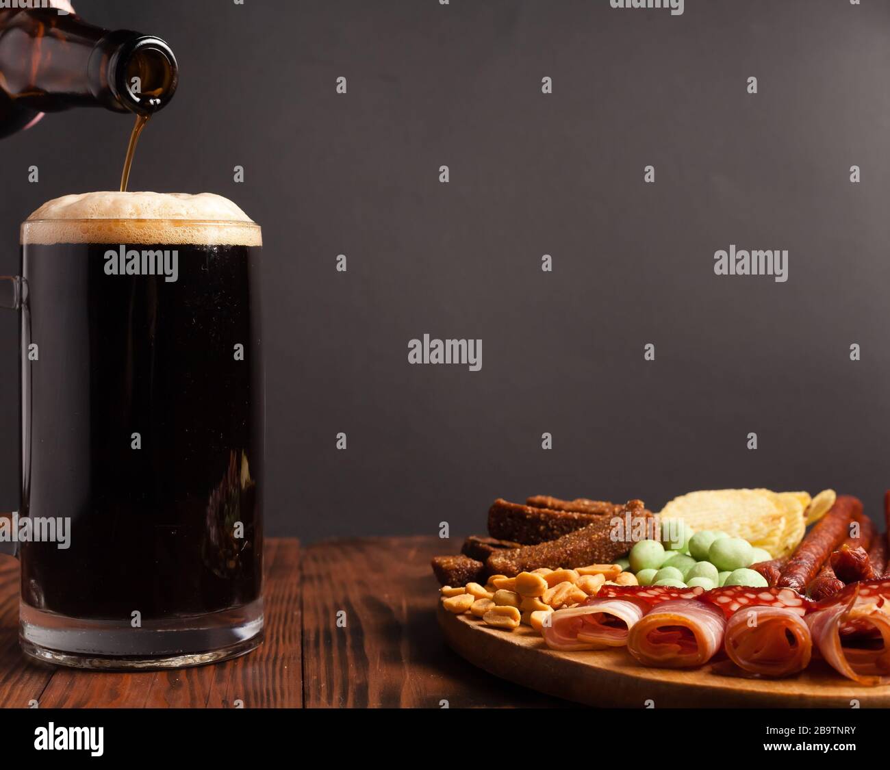 Tazza di birra e assortimento di spuntini su un tavolo di legno. Salsiccia, salame, prosciutto, pancetta. Snack salato alla birra, patatine, noci wasabi, arachidi salate, crostini. Foto Stock