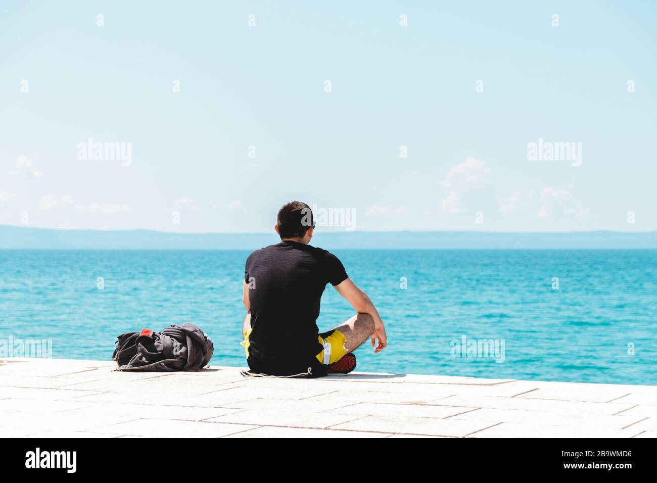 giovane uomo viaggiatore solo con zaino vicino a lui meditando guardando l'oceano e godendo il silenzio Foto Stock