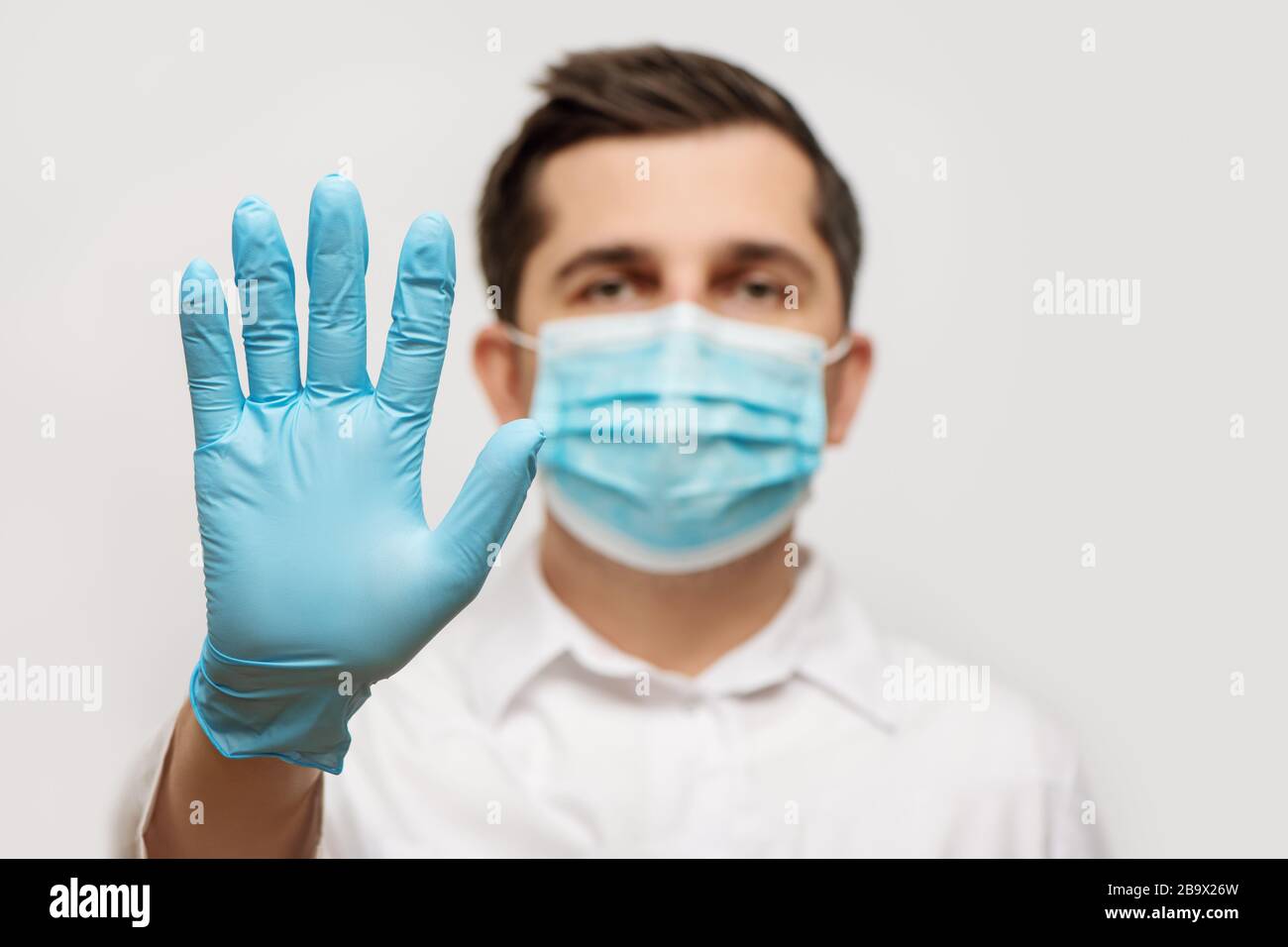 Ritratto del medico in uniforme medica con maschera facciale protettiva blu e una mano con guanti che mostra un segnale di stop. Interrompere il concetto COVID-19. Foto Stock