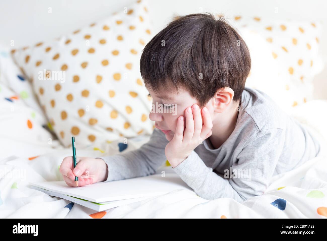 Ragazzo della scuola a letto e fare i compiti della scuola a casa, scrivendo in quaderno. Formazione online a distanza. Quarantena Foto Stock