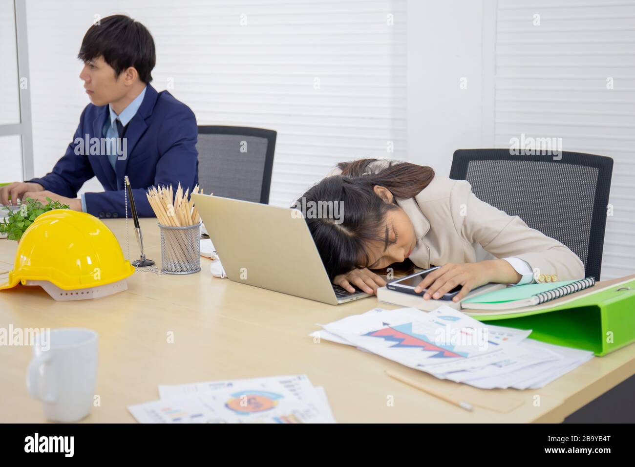 Una giovane e professionale donna asiatica d'affari dorme sulla sua scrivania e un dipendente di sesso maschile sta lavorando. Foto Stock