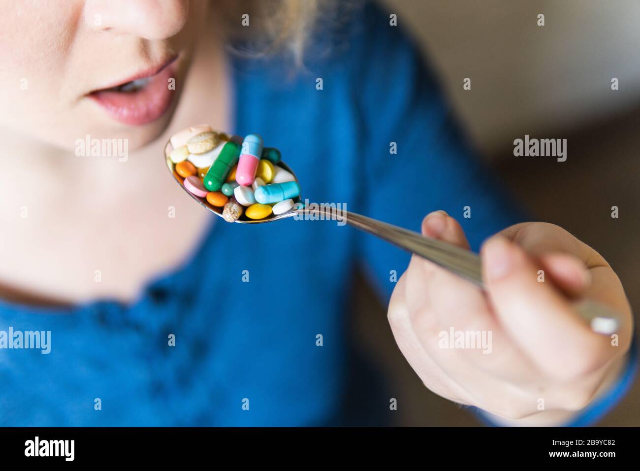 Giovane donna bionda che tiene un cucchiaio pieno di pillole assortite davanti alla sua bocca aperta. Foto Stock