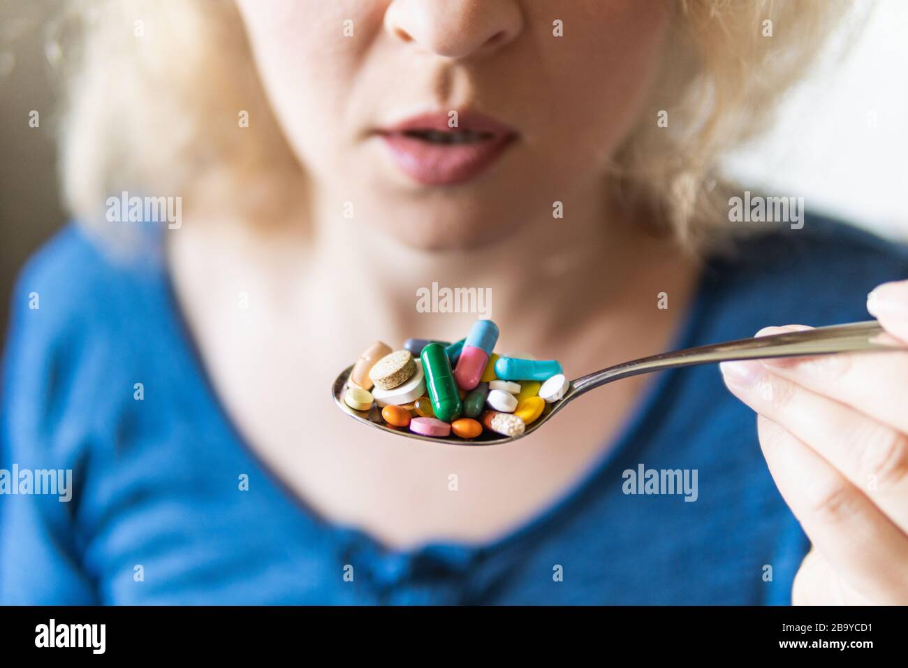 Giovane donna bionda che tiene un cucchiaio pieno di pillole assortite davanti alla sua bocca aperta. Foto Stock