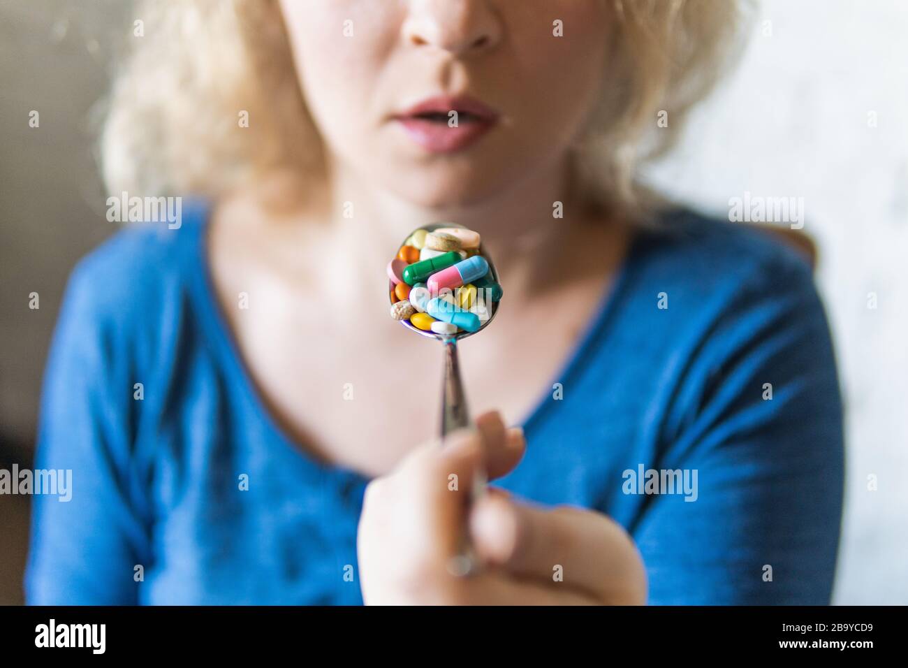 Giovane donna bionda che tiene un cucchiaio pieno di pillole assortite davanti alla sua bocca aperta. Foto Stock