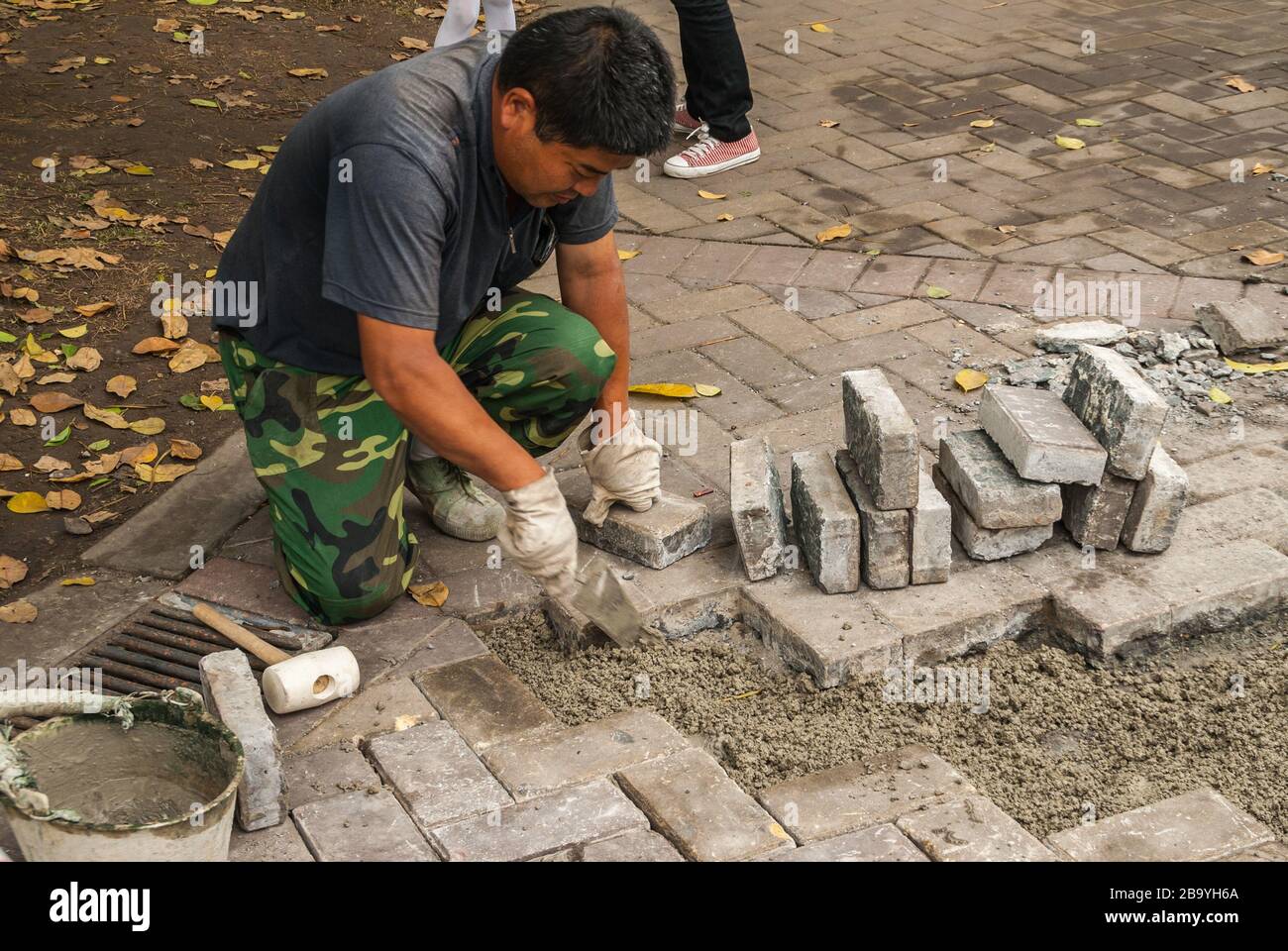 Chongqing, Cina - 9 maggio 2010: Lavoratore maschio in pantaloni camouflage verde e camicia grigia mette manualmente la superficie del marciapiede insieme con mattoni grigi. Foto Stock