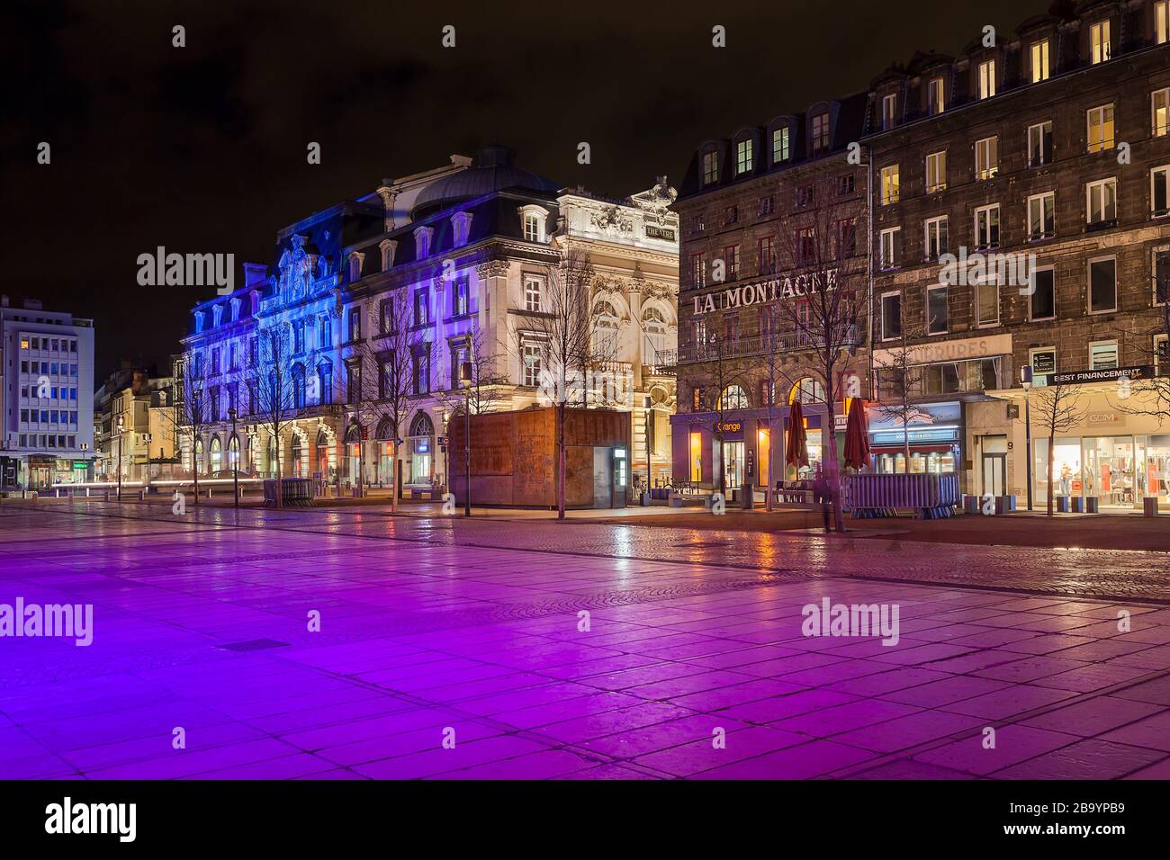 Vista notturna di piazza Jaude (Place de Jaude), Clermont Ferrand in Francia, illuminata da luce viola, che circonda tradizionali edifici francesi e negozi Foto Stock