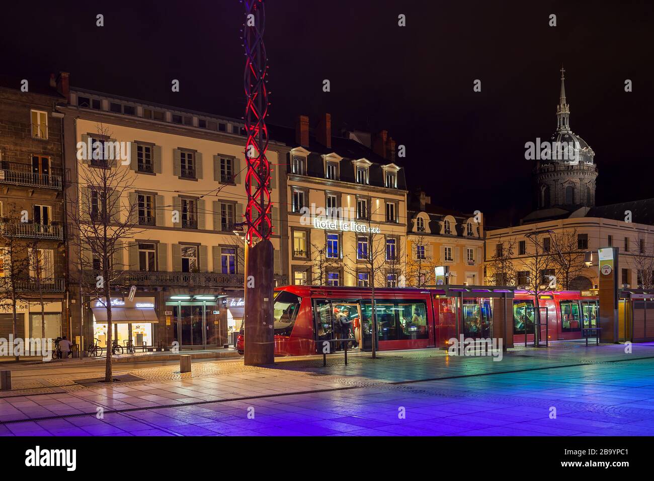 Vista notturna di Place de Jaude a Clermont Ferrand, Francia, con tram rosso sulla stazione, luci della città e chiesa di Saint Pierre des Minimes Foto Stock
