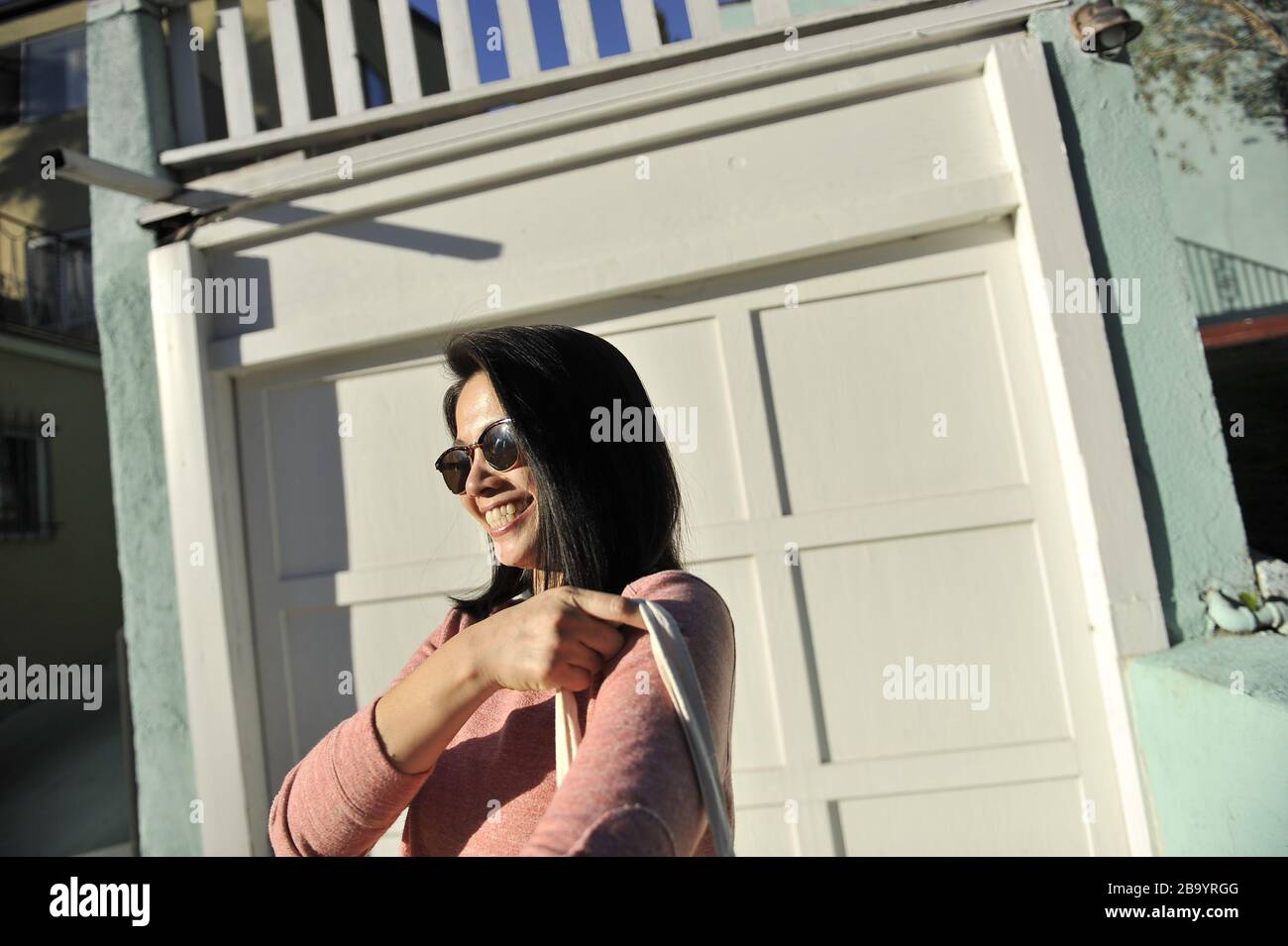 Donna asiatica in maglione rosa e occhiali da sole sorridenti candida all'esterno Foto Stock