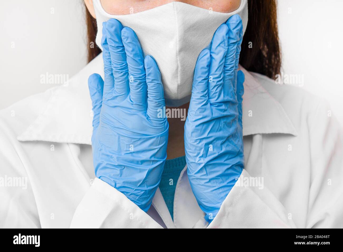 Closeup donne mani al viso in maschera protettiva contro coronavirus, SARS-cov-2, 2019-nCoV o influenza. Medico in camice bianco, guanti blu e maschera. Foto Stock