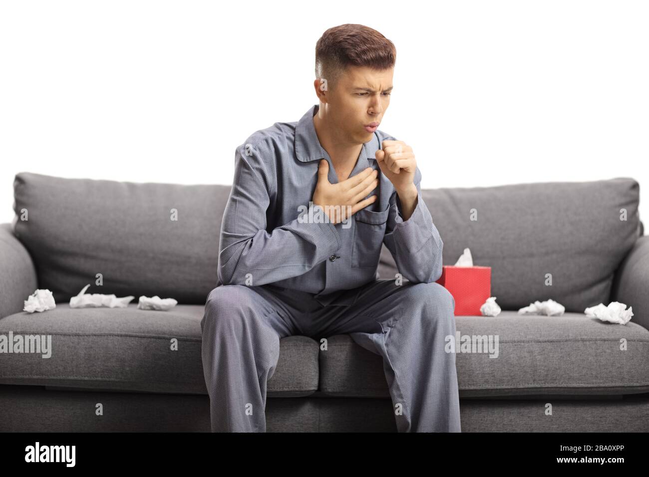 Giovane uomo tosse e seduto su un divano in pigiama con carte di tessuto isolato su sfondo bianco Foto Stock