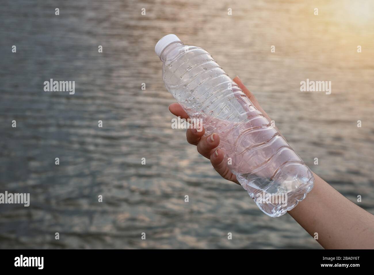 Chiudi la mano della donna tenendo la bottiglia di plastica trasparente sullo sfondo del fiume Foto Stock