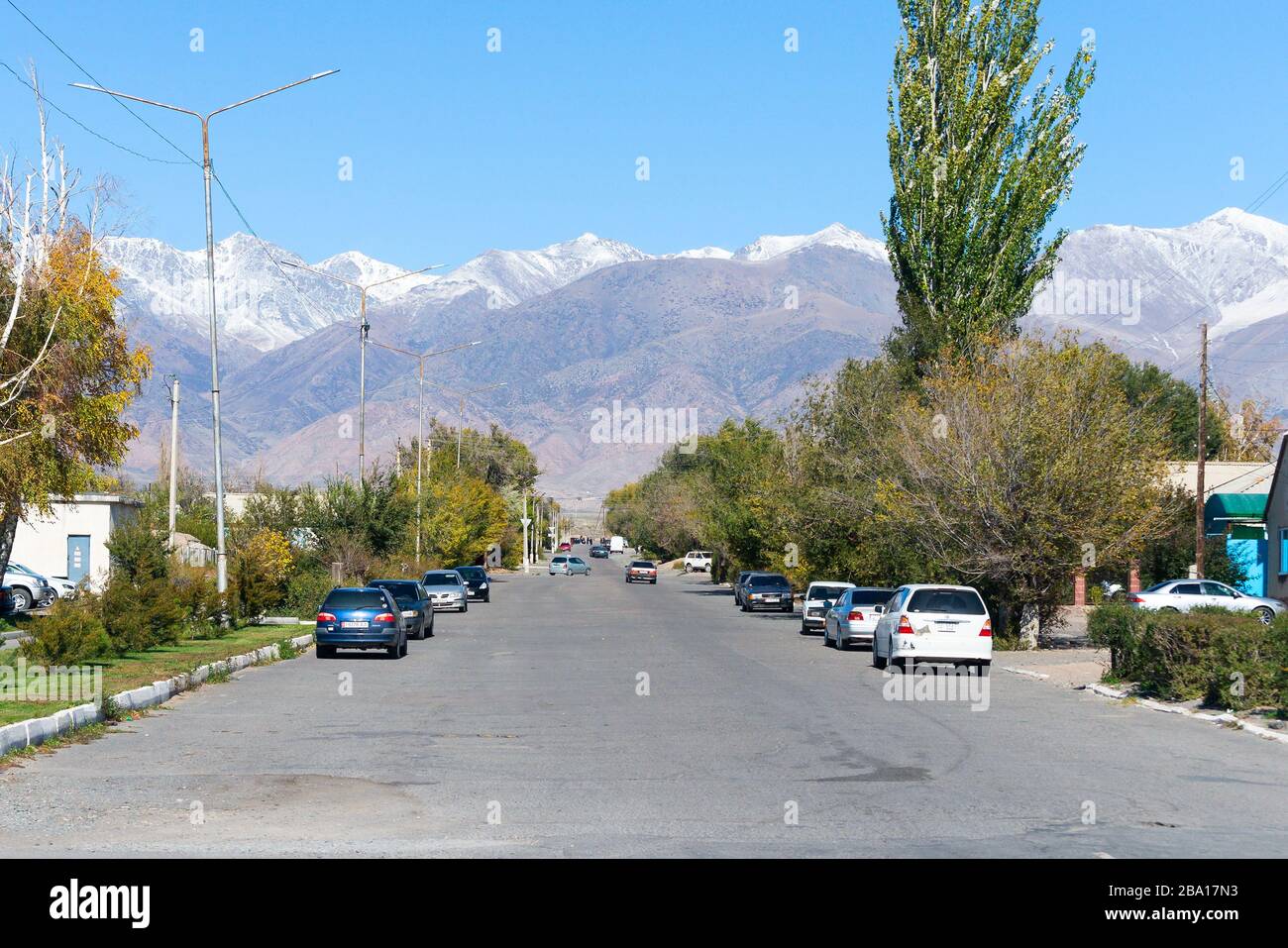 Strada asfaltata con auto parcheggiate in una piccola città vicino al lago Issyk Kul. Montagne ricoperte di neve e alberi visibili a Balykchy, Kirghizistan. Foto Stock