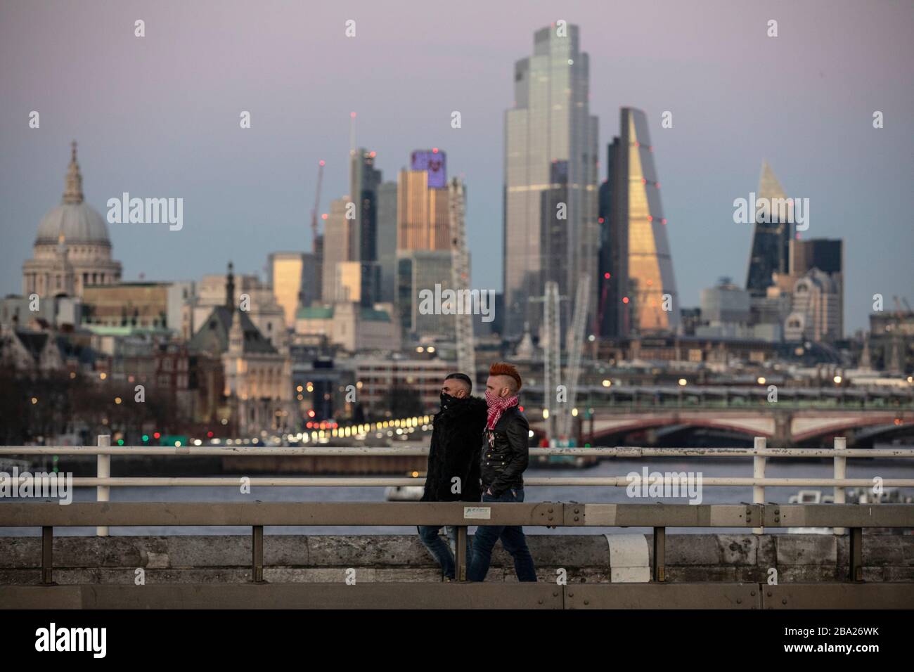 Londra, Regno Unito. 25 Marzo 2020. Due uomini camminano attraverso il Waterloo Bridge proteggendo le loro bocche dall'infezione di Covid-19 con la City of London in lontananza, come gli economi prevedono che un incidente potrebbe costare più vita di Cronavirus. Credit: Jeff Gilbert/Alamy Live News Foto Stock