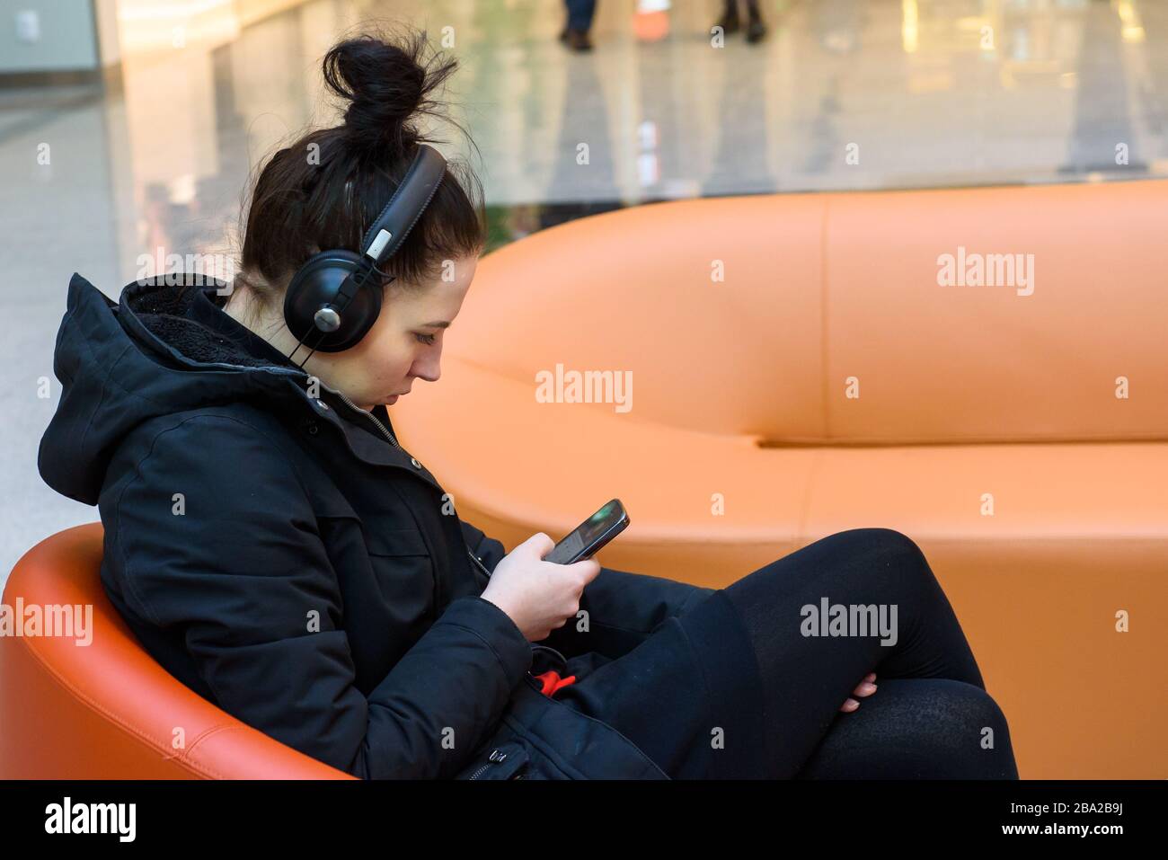 RIGA, LETTONIA. 4 aprile 2019. Giovane donna ascolta la musica e legge il messaggio sullo smartphone. Seduta sulla poltrona. Foto Stock