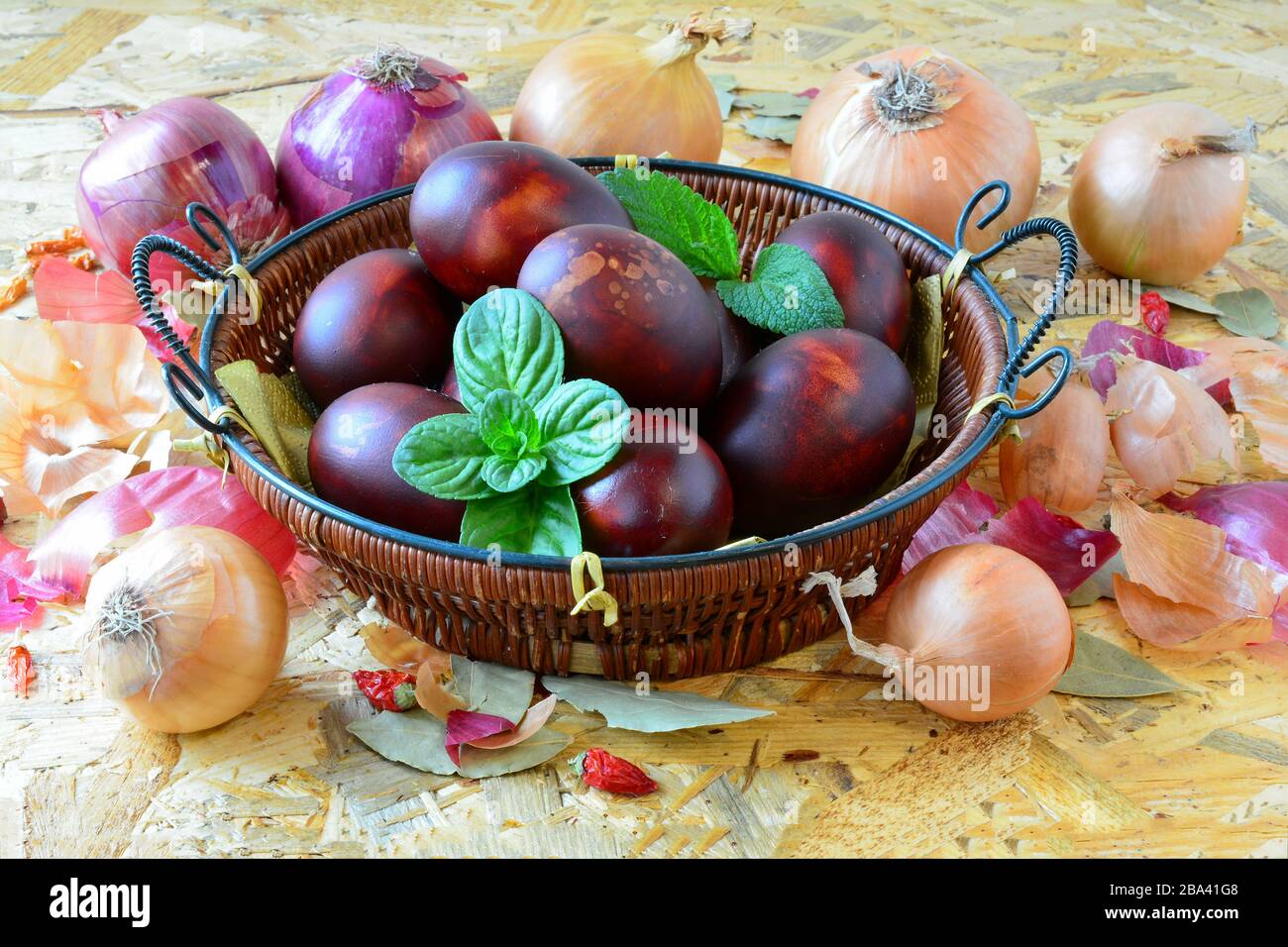 Uova di Pasqua in cesta di vimini e cipolla gialla e rossa, peperoni e basilico verde e foglie di balsamo al limone su sfondo di legno, primo piano Foto Stock