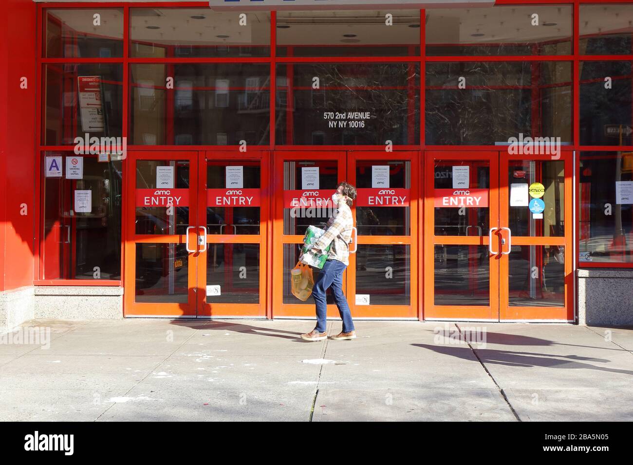 New York, NY, 24 marzo 2020. Sullo sfondo di un cinema AMC chiuso, una persona che indossa una maschera N95 e guanti usa e getta porta custodie di acqua in bottiglia e un sacchetto di carta igienica durante la pandemia COVID-19 coronavirus. Credito: Robert K. Chin. Foto Stock