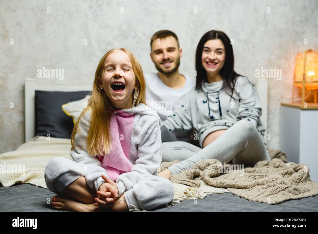 Affascinante ragazza ridente con lunghi capelli biondi a letto in pijamas con genitori sullo sfondo Foto Stock