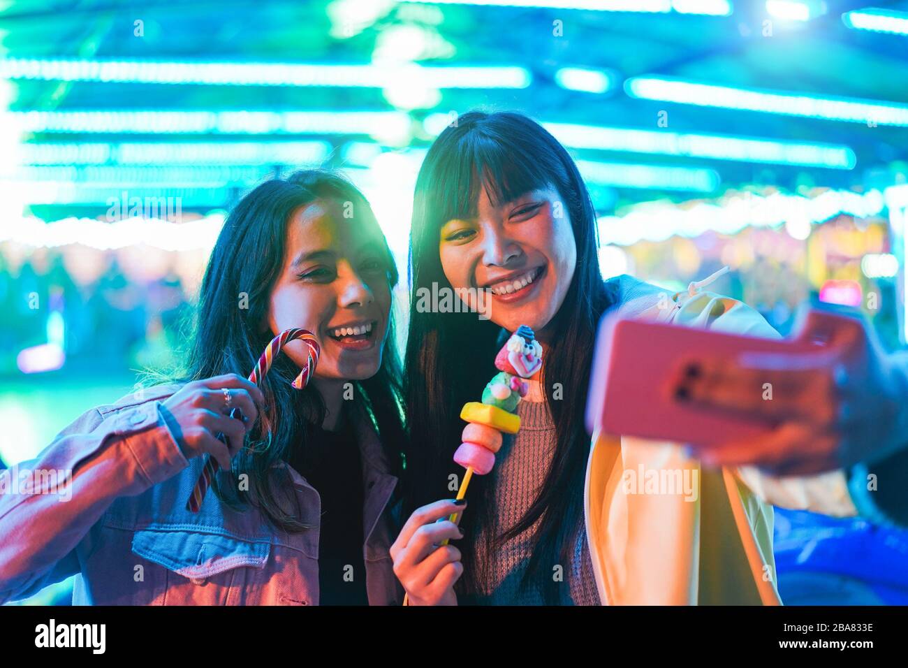 Ragazze asiatiche felici mangiare caramelle e prendere selfie al parco divertimenti - giovani amici alla moda divertirsi con la tendenza della tecnologia - tecnologia, amicizia A. Foto Stock