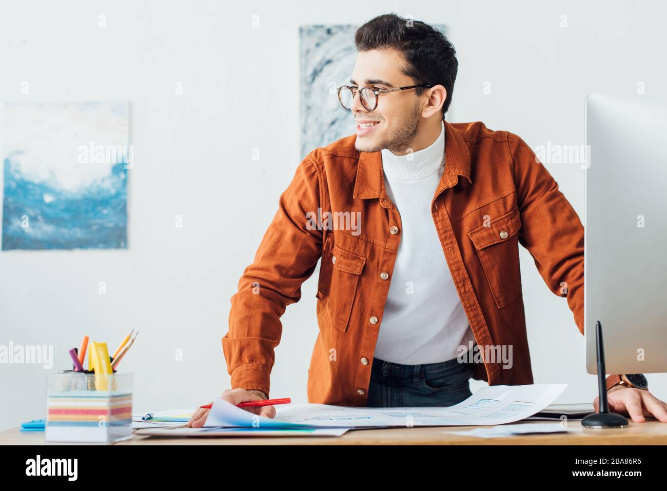Bel progettista ux sorridente mentre lavora con i modelli vicino al computer al tavolo in ufficio Foto Stock