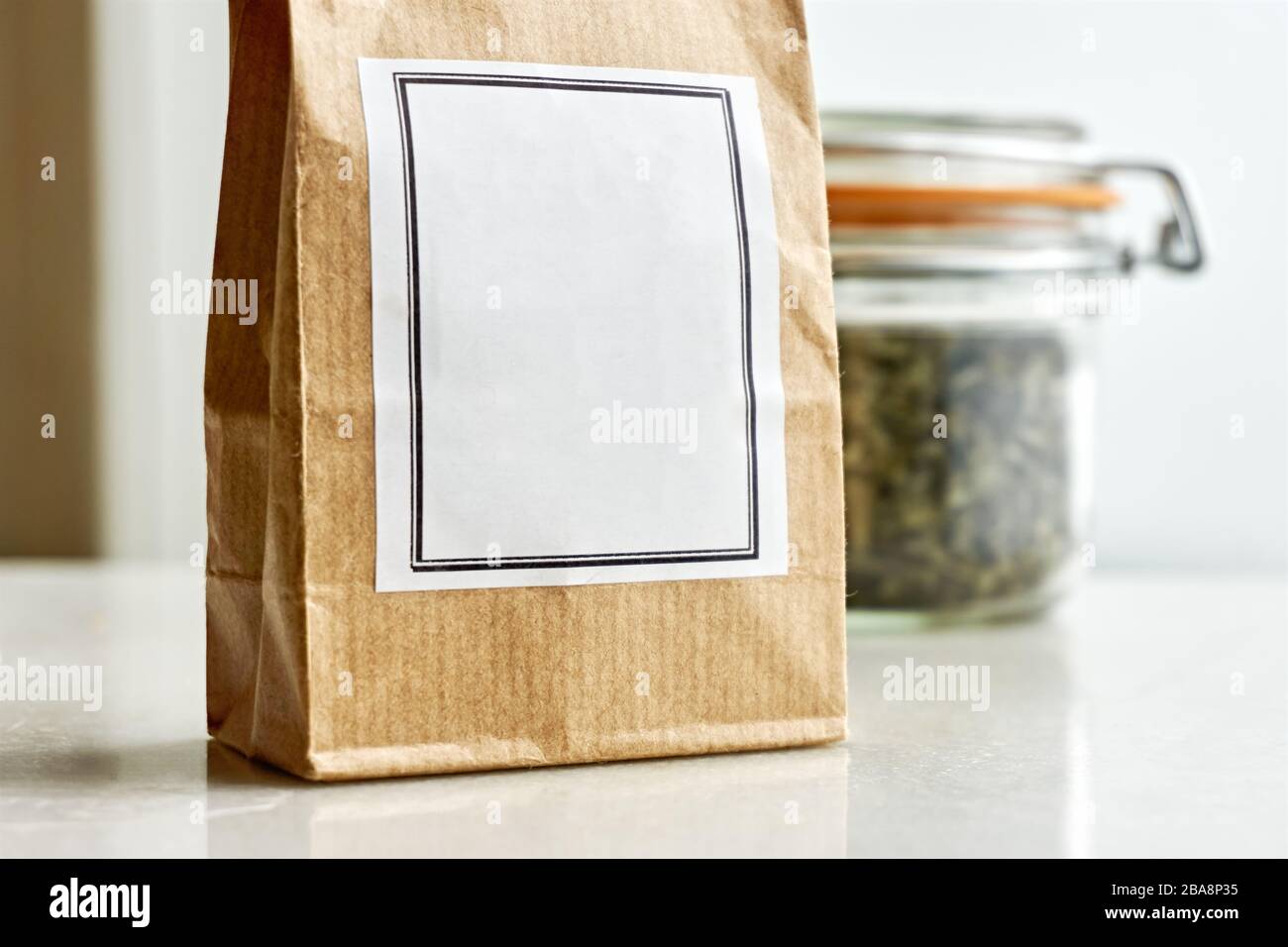 Sacchetto di carta marrone kraft con etichetta bianca bianca davanti a un vaso di vetro della balla e guarnizione di gomma arancione con erbe essiccate o tè su uno sfondo bianco. Foto Stock