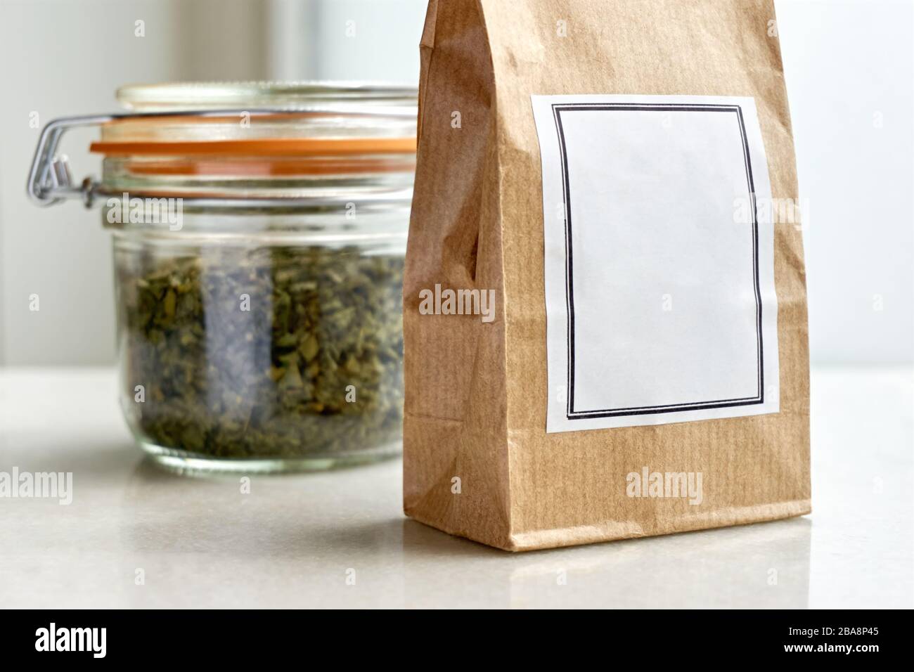 Sacchetto di carta marrone kraft con etichetta bianca bianca davanti a un vaso di vetro della balla e guarnizione di gomma arancione con erbe essiccate o tè su uno sfondo bianco. Foto Stock