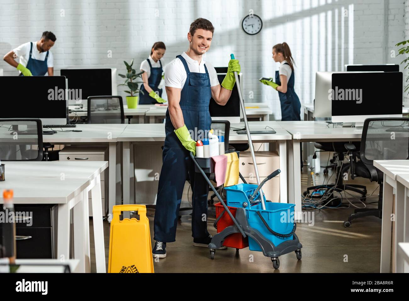 allegro pulitore in movimento carrello con forniture di pulizia vicino a colleghi multiculturali Foto Stock