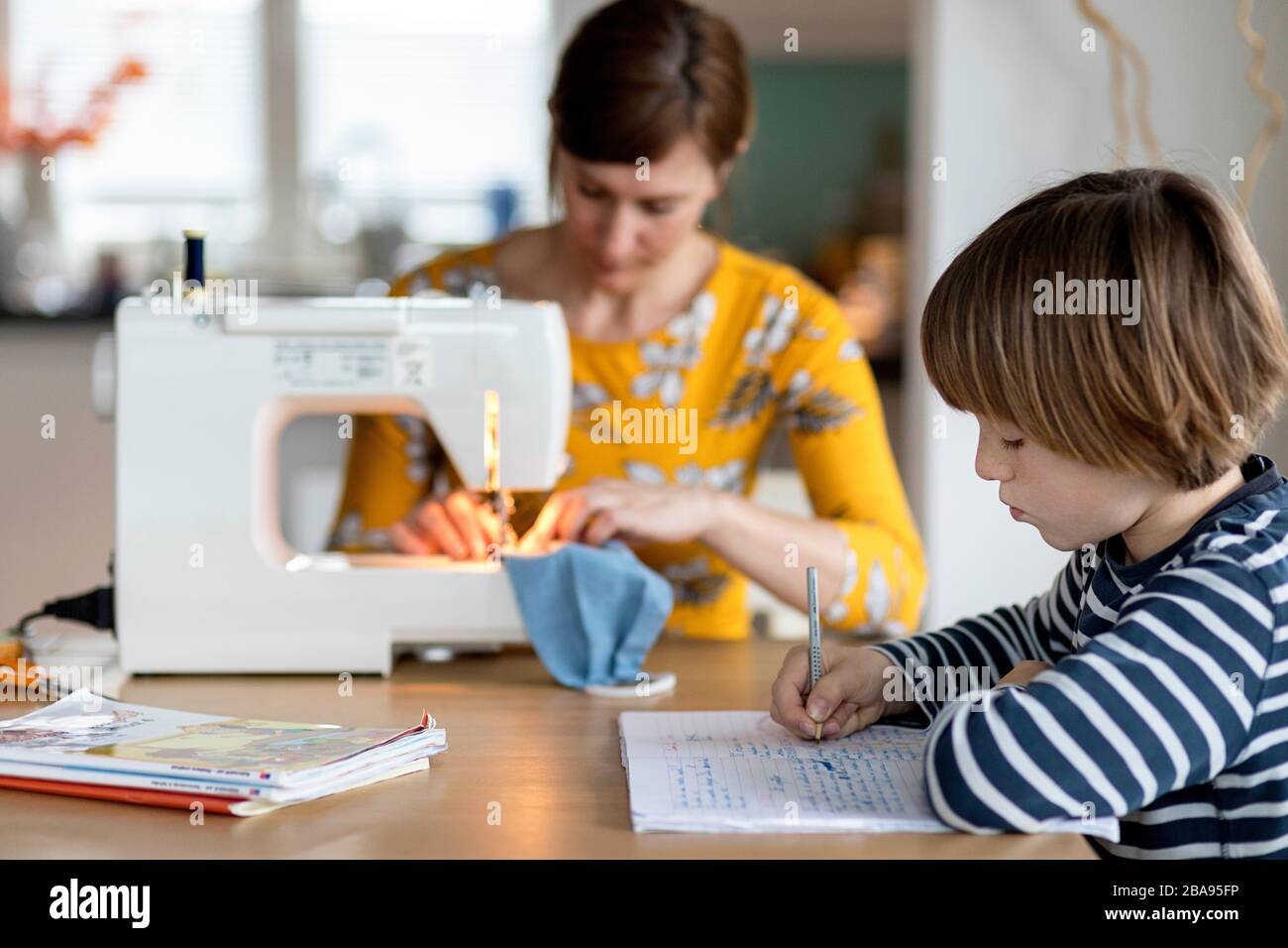 Donna che fa la maschera del viso fatta in casa con una macchina da cucire, bambino homeschooling a casa mentre in quarantena, protezione contro il virus corona, marzo 2020 Foto Stock