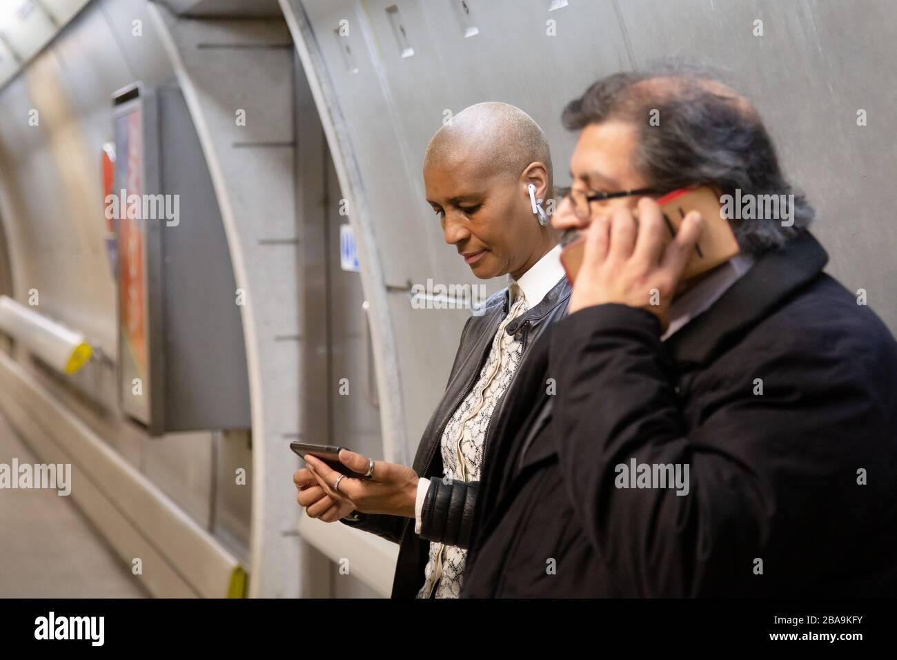 Una donna usa il suo smartphone sulla metropolitana mentre un uomo effettua una chiamata telefonica, parte del sistema della metropolitana di Londra Foto Stock