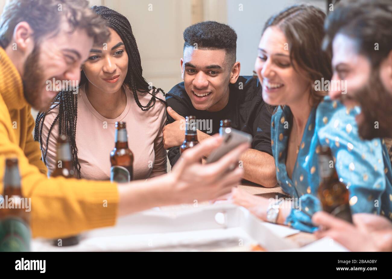 Gruppo di giovani sorridenti che guardano video su smartphone e bevono birre in una casa. Amici multirazziali che si divertono con lo smartphone, social netw Foto Stock