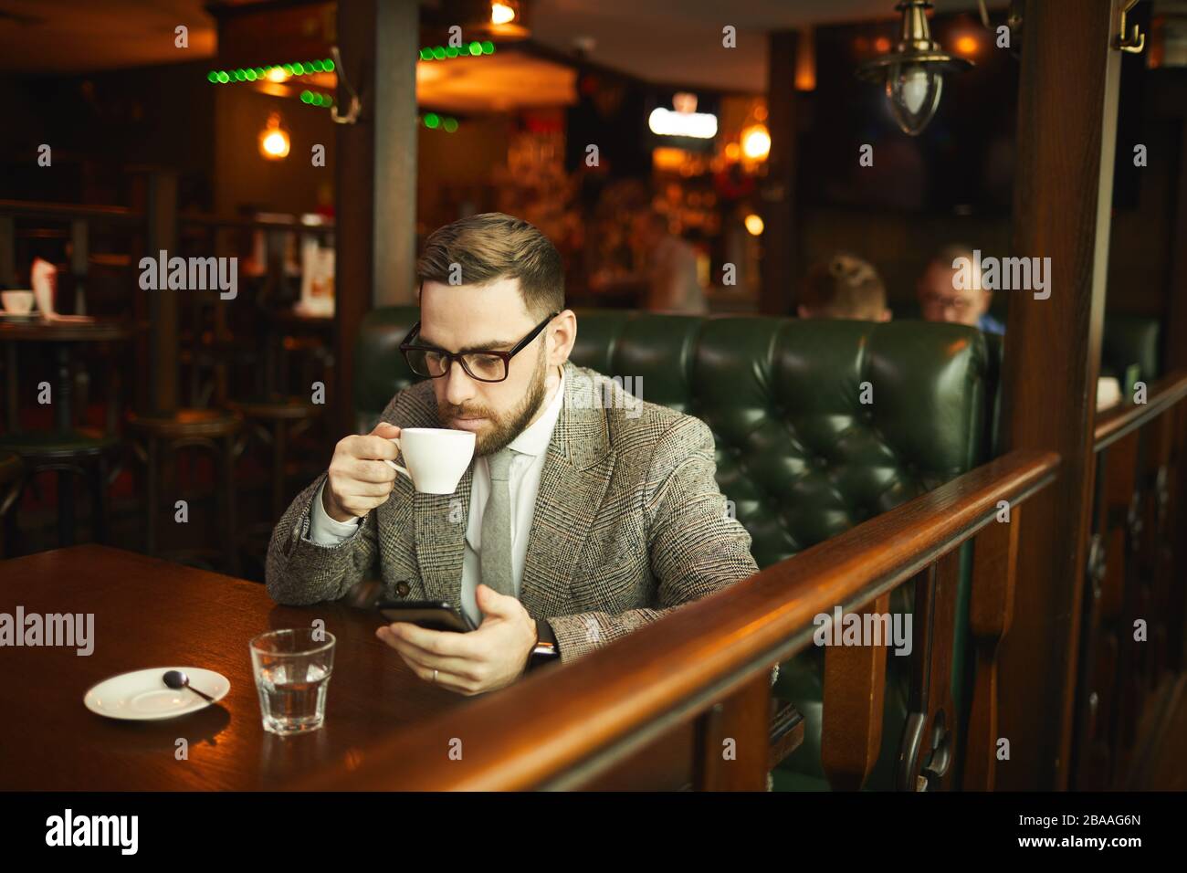 Giovane uomo d'affari in occhiali e in tuta bere caffè e utilizzando il suo telefono cellulare mentre si siede al tavolo in bar Foto Stock