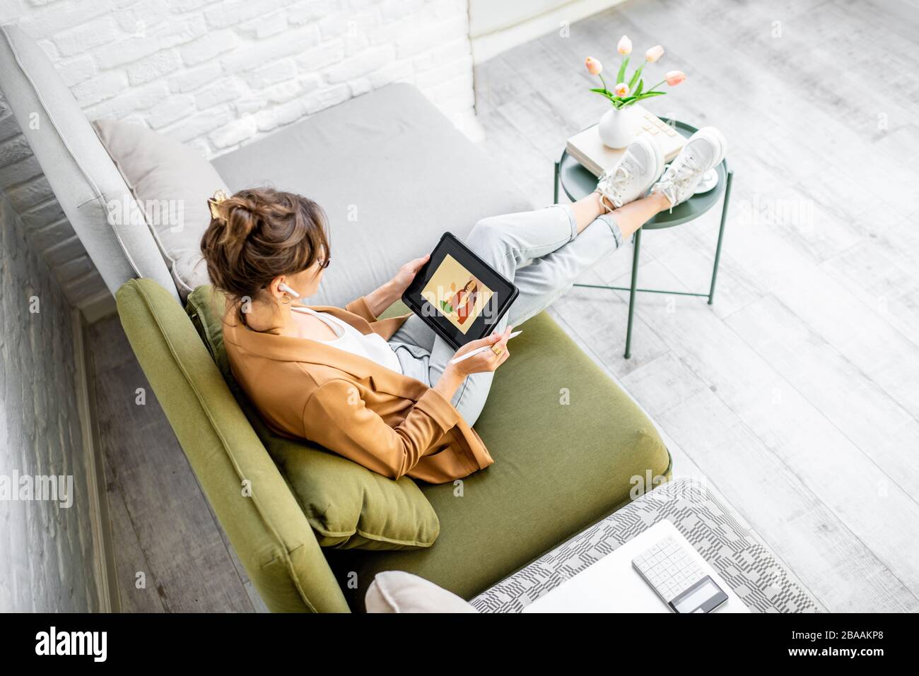 Giovane donna creativa che lavora come web designer o artista, disegnando su un tablet digitale mentre si siede sul comodo divano a casa Foto Stock