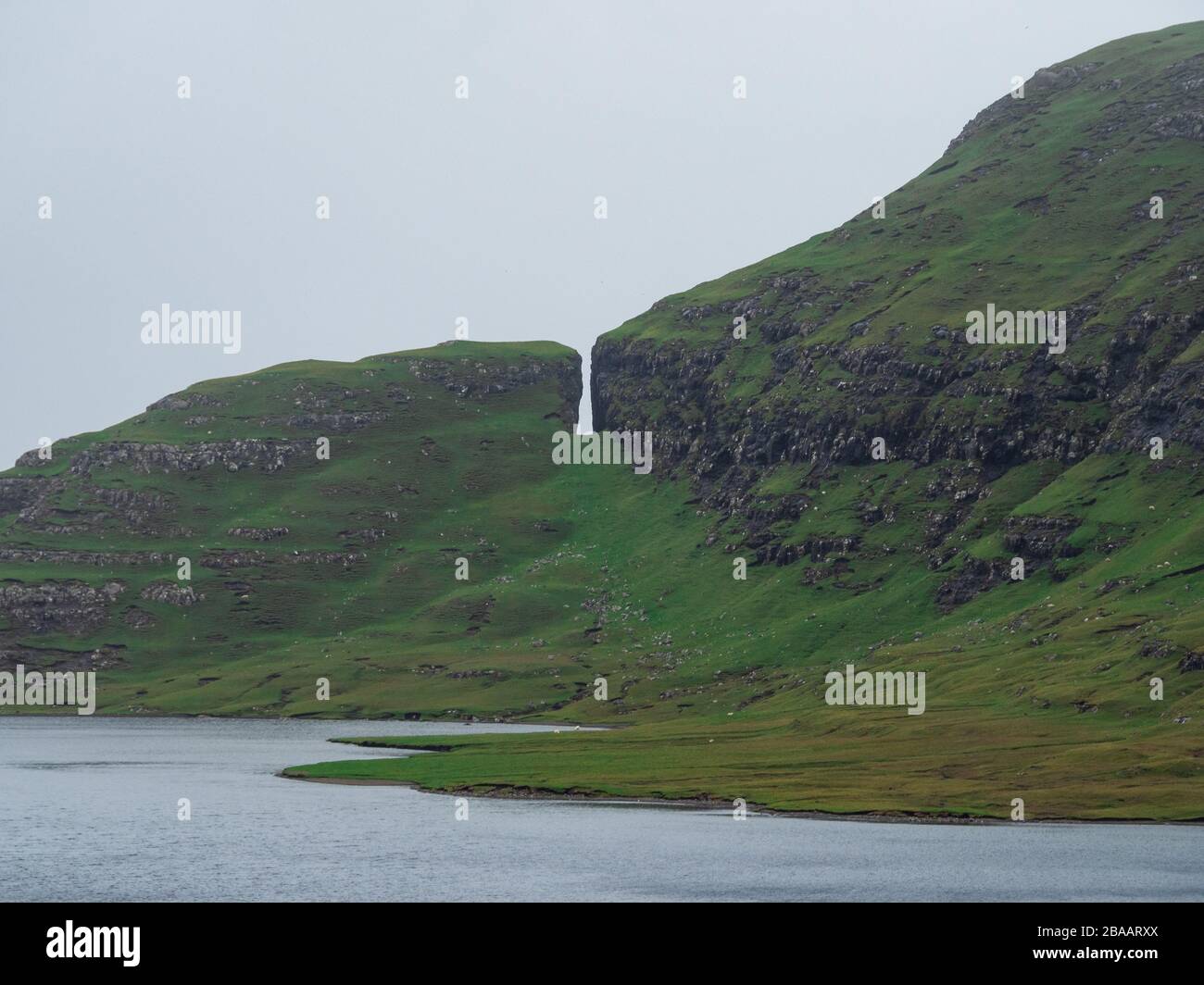 Verdi colline e scogliere delle Isole Faroe. Lago Leitisvatn. Strana formazione rocciosa. Tempo frizzante. Foto Stock