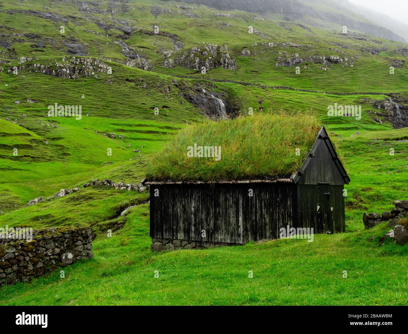 Isole Faroe. Saksun. Vecchia casa in legno con tetto in erba. Campi verdi, cascata sul villaggio. Paesaggio frizzante. Nuvole sulle colline. Foto Stock