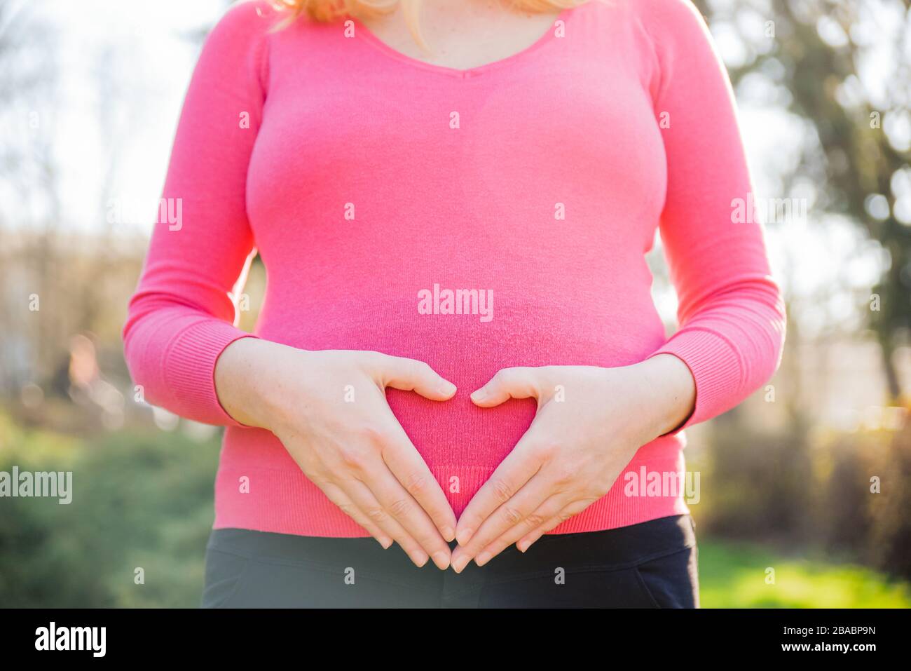 Donna incinta in camicia rosa nel mezzo di una cornice in piedi fuori su un pomeriggio soleggiato di primavera e formare un segno di un cuore sul suo pancino Foto Stock