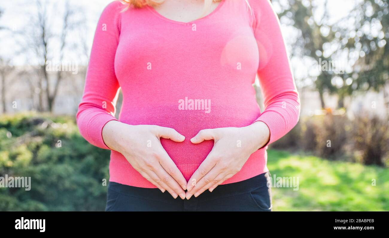 Donna incinta bionda in piedi fuori su una soleggiata primavera pomeriggio e tenendo le mani sul suo ventre Foto Stock