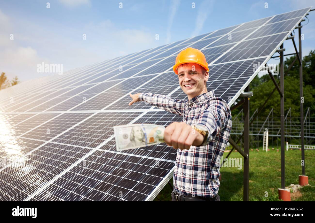 Ragazzo sorridente con cento dollari di legge in mano mostra alla stazione di pannelli solari in cui i raggi del sole sono riflessi. Concetto di business e generazione. Generazione di energia ecologica. Foto Stock