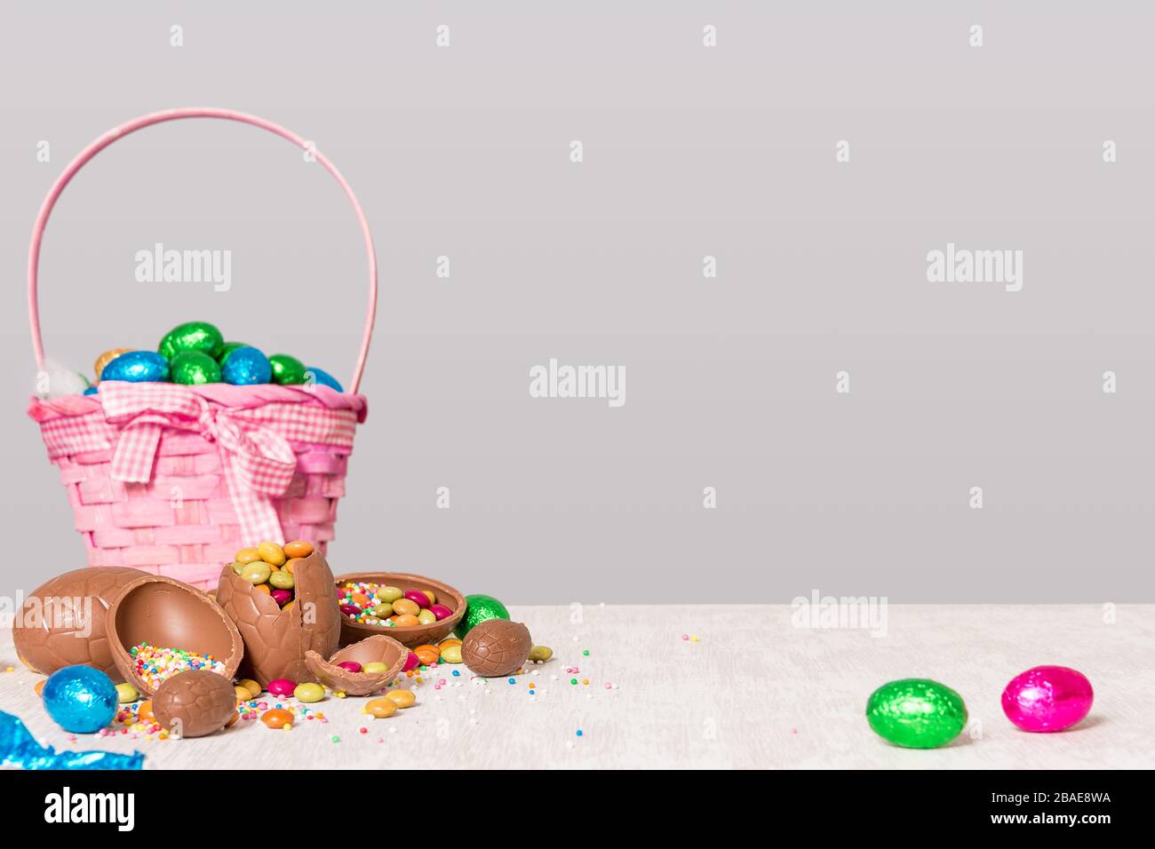 cioccolato uova di pasqua in un cestino e su tavolo di legno con spruzzette multicolore e dolci che sparge fuori sul tavolo - concetto di festa di pasqua Foto Stock