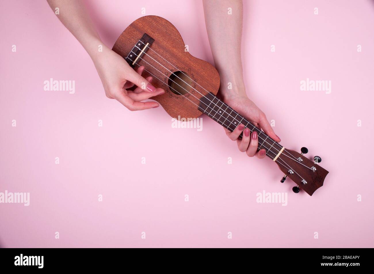 Donna mani con bellezza rosa manicure tenendo piccolo wodden ukulele su sfondo rosa. Bella foto di musicista femminile. Foto Stock