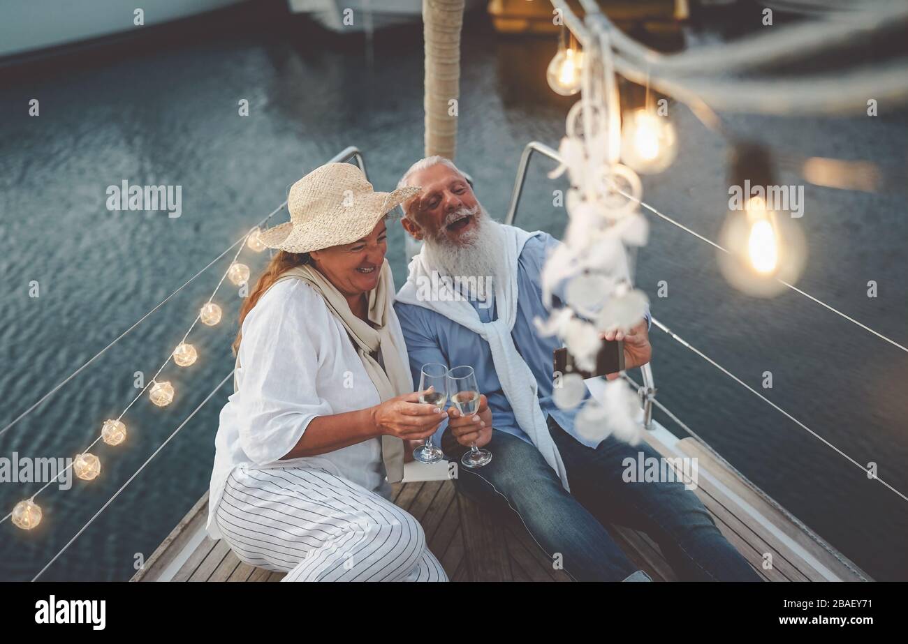 Coppia senior che brindano champagne mentre si prende selfie in vacanza in barca a vela - gente matura felice che si divertono a celebrare l'anniversario di matrimonio in barca Foto Stock
