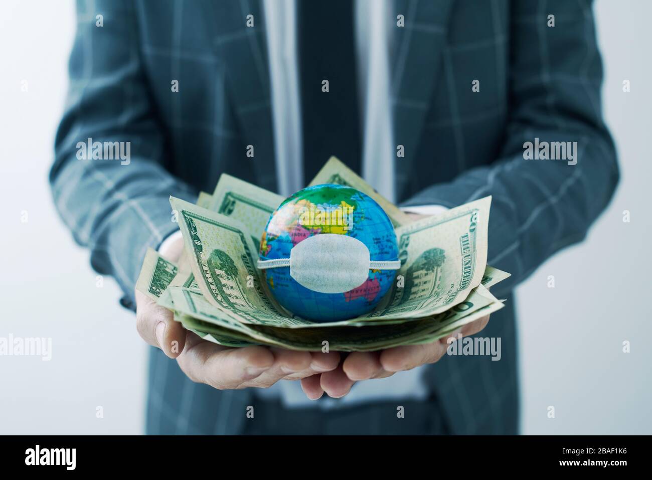 uomo d'affari che detiene un globo mondiale con una maschera protettiva in cima a una manciata di dollari, che raffigura le conseguenze economiche del coronavirus p Foto Stock