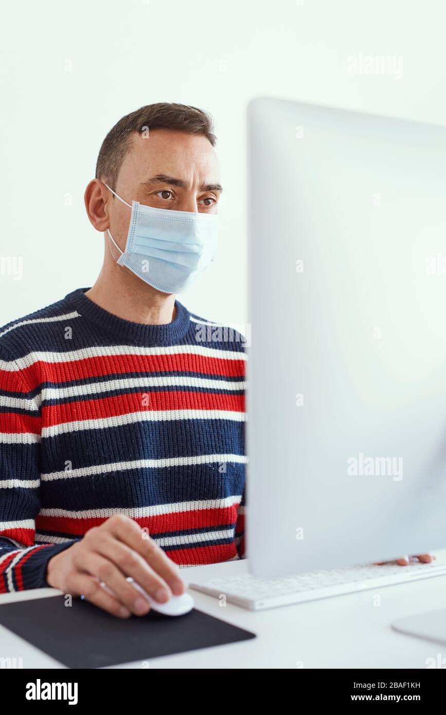primo piano di un giovane caucasico all'interno, con una maschera chirurgica, con un computer seduto su un tavolo bianco Foto Stock